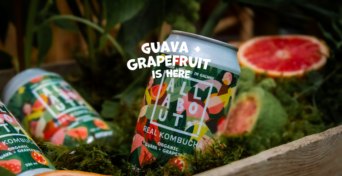 A can of All About Kombucha Real Kombucha in Guava + Grapefruit flavor is displayed in a wooden tray filled with moss, alongside sliced grapefruit and other cans, with a text overlay reading 'GUAVA + GRAPEFRUIT IS HERE'.