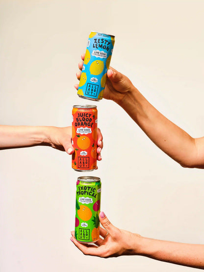Three hands hold three colorful beverage cans stacked vertically against a plain light beige background. The cans are labeled Zest Lemon, Juicy Blood Orange, and Exotic Tropical, with bright fruit illustrations and bold text.